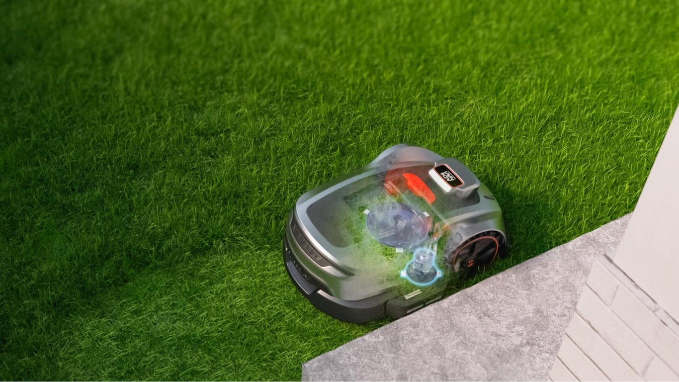 Robotplæneklipper kører tæt på kantsten ved hus med delvist gennemsigtig visning af interne mekaniske dele.