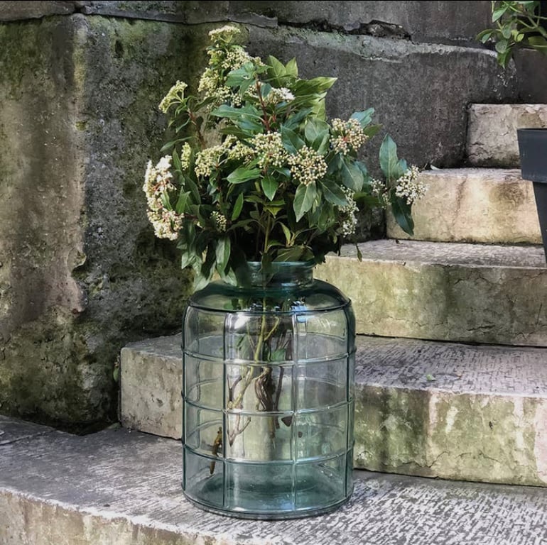 Glasvase med grønt løv og hvide blomster placeret på betontrappe med mosgroet væg i baggrunden. (2)