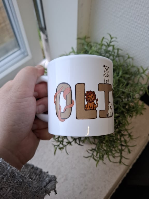 Hånd holder en hvid kop med tegninger af dyr og bogstaverne "OLI" med planter og et vindue i baggrunden. (4)