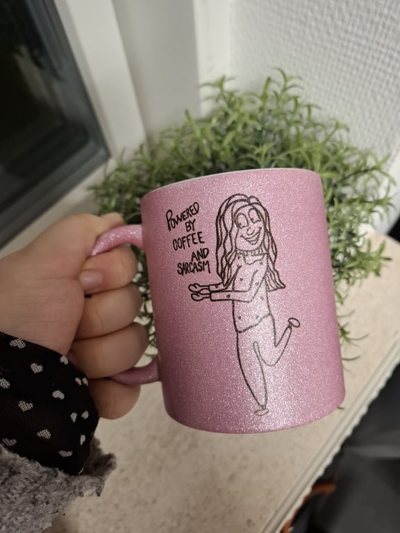 Hånd holder glitrende lyserød kop med tegning af en kvinde og teksten "Powered by coffee and sarcasm" foran en grøn plante.