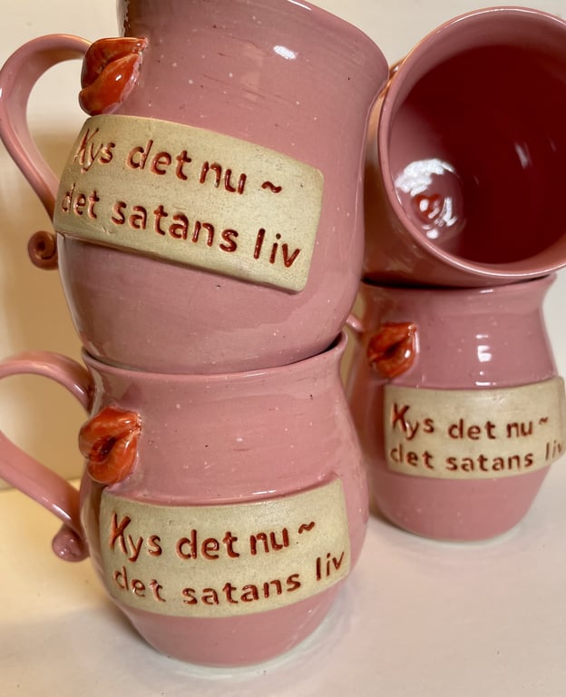 Tre rosa keramikkrus med teksten "Kys det nu ~ det satans liv" samt en lille læberfigur ved hankene.