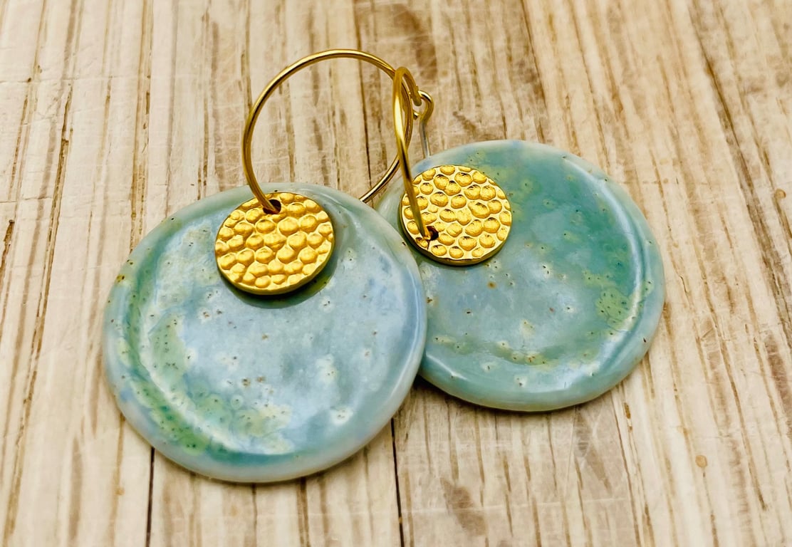 Nærbillede af runde øreringe med blågrønne keramiske skiver og små, runde, hamrede gulddetaljer på en lys træbaggrund.