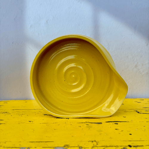 Gul keramikskål med spiralformet mønster, placeret på et gulmalet bord med hvid væg i baggrunden. (1)