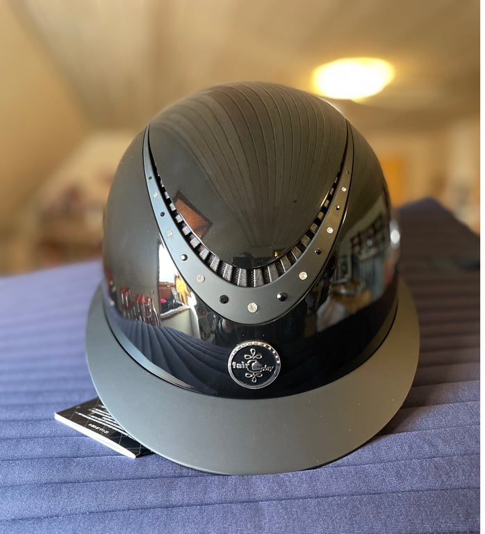 Sort blank ridehjelm med ventilationsåbninger og smykkeagtige detaljer, placeret på en blå overflade indendørs.