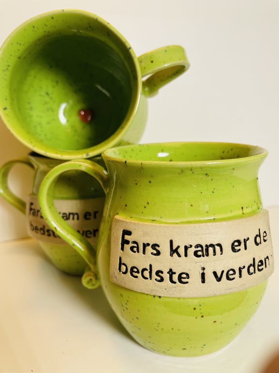 To grønne krus med tekst "Fars kram er de bedste i verden" på en lys baggrund.