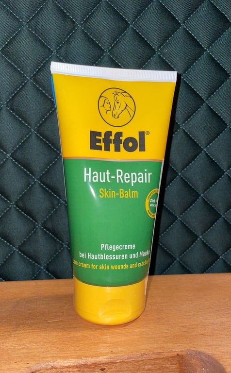 Tube med Effol Haut-Repair Skin-Balm, en plejecreme til hudsår og sprækker, placeret på træbord foran en quiltet mørk baggrund.