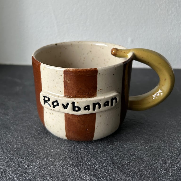 Keramikkrus med brun og beige stribet mønster, med håndtag formet som en gul banan, teksten "Røvbanan" skrevet på fronten.