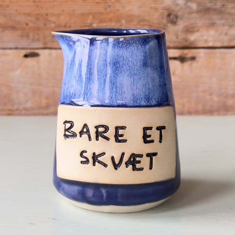 Keramisk mælkekande med blå glasur og teksten "Bare et skvæt" på en beige label.