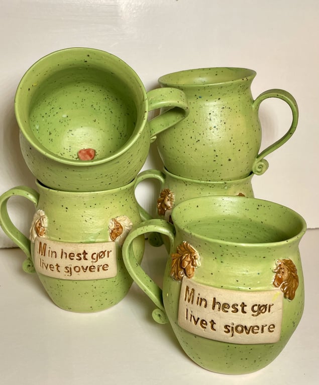 Grønne keramikkrus med tekst på dansk "Min hest gør livet sjovere" og hestehoveder som dekorative detaljer.