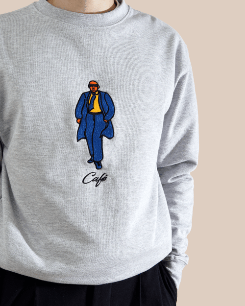 Person iført grå sweatshirt med broderet illustration af en mand i blå frakke og bukser, gul skjorte og solbriller, samt teksten "Café" under. (1)