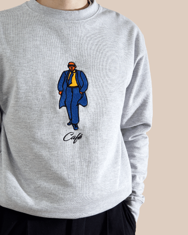 Person iført grå sweatshirt med broderet illustration af en mand i blå frakke og bukser, gul skjorte og solbriller, samt teksten "Café" under.