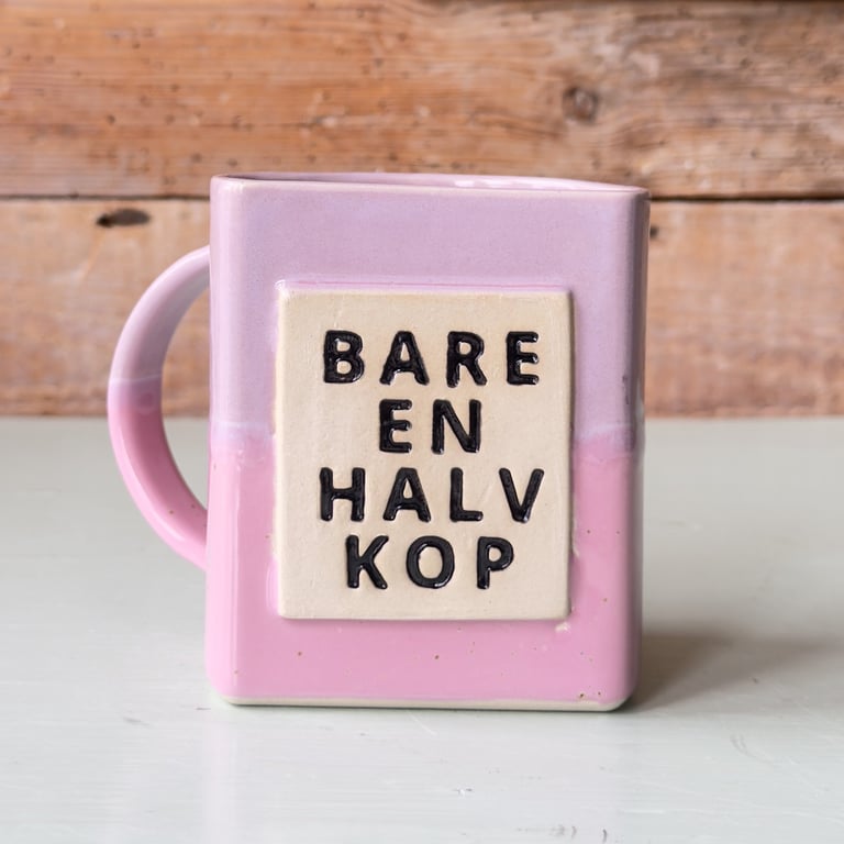 Lyserød keramikkop med firkantet form og tekst "BARE EN HALV KOP" i sort skrift på fronten.