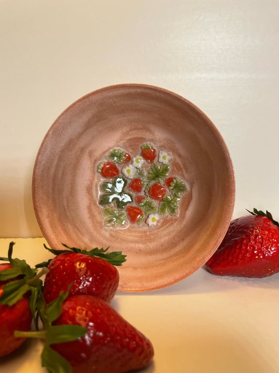 Lille rosa keramikskål med glasurmaleri af jordbær og blomster, omgivet af friske jordbær.