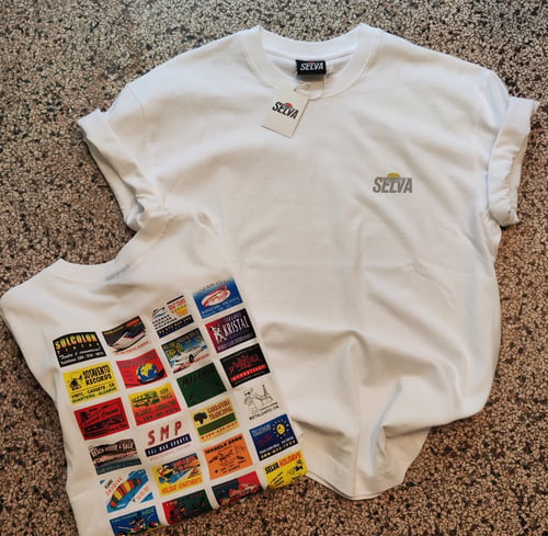 To hvide t-shirts med logoet "SELVA"; den ene med tryk foran og den anden med farverige, nostalgiske reklamer på ryggen, lagt på en stenbelægning. (1)
