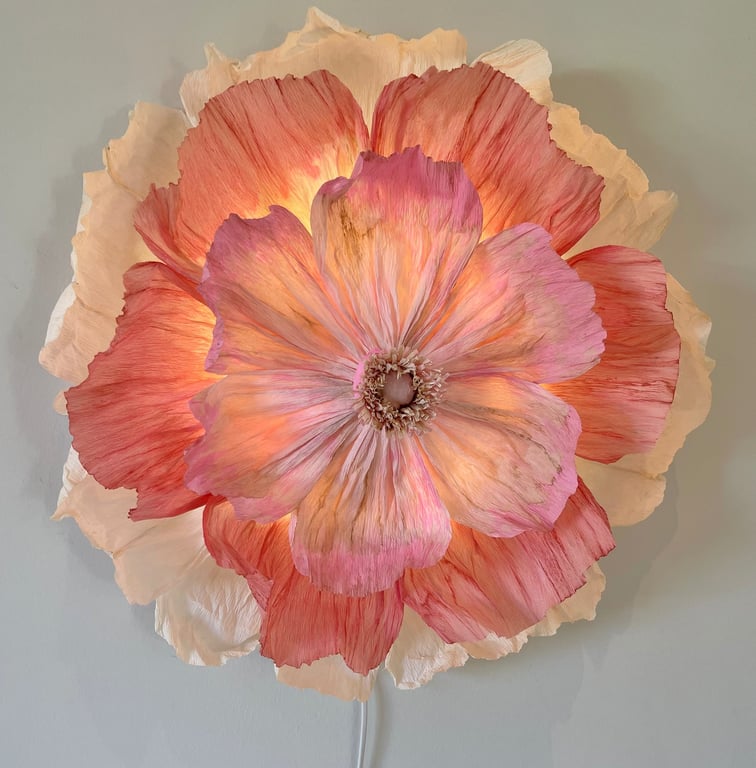 Stor væglampe formet som en blomst med rosenrøde og cremefarvede kronblade, oplyst indefra.
