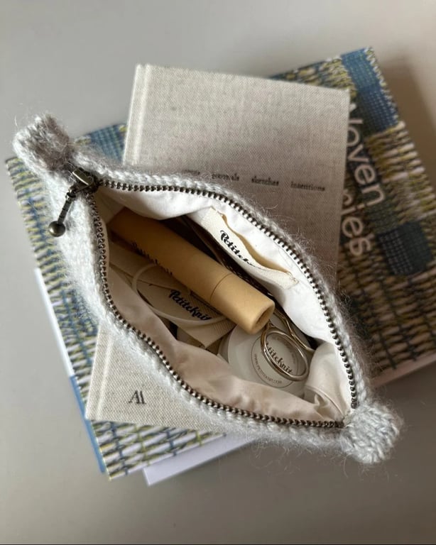 En åben strikketaske med lynlås, der indeholder strikkeværktøj som nøglestrimler og låse-maskemarkører, placeret ovenpå to bøger.