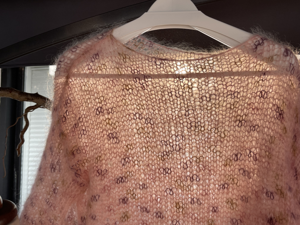 Lyserød, let og luftig mohairstriktrøje med mønster, hængende på en hvid bøjle foran et vindue med dagslys bagved. (3)