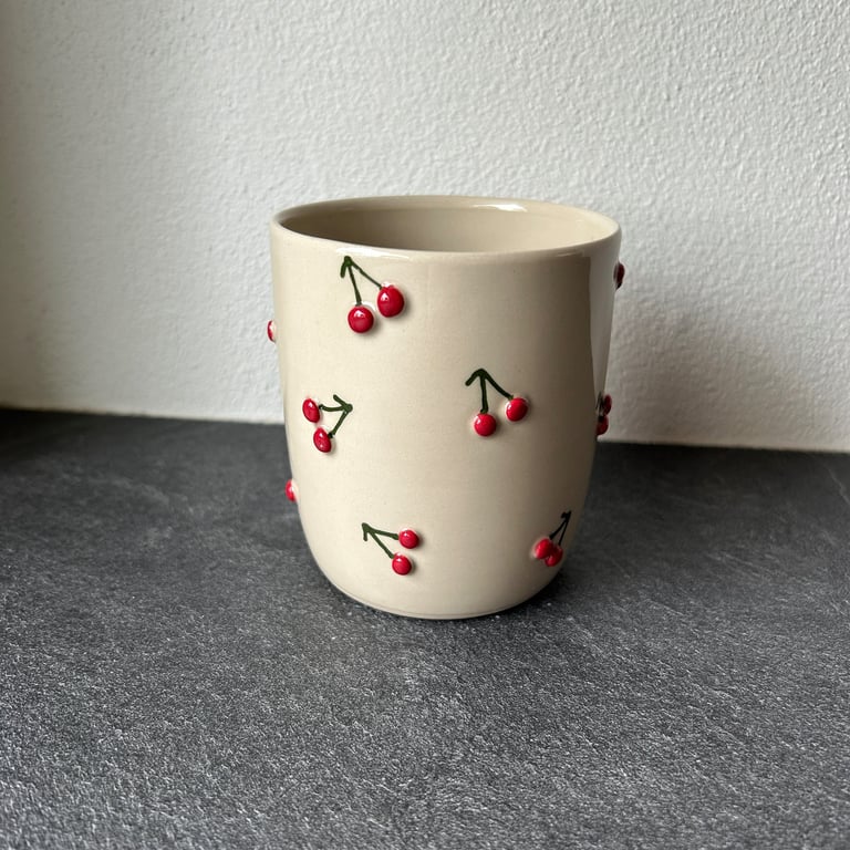 Kræmmerhuset kop i beige med små røde kirsebær og grønne stilke som pynt, står på gråt bord mod hvid væg.