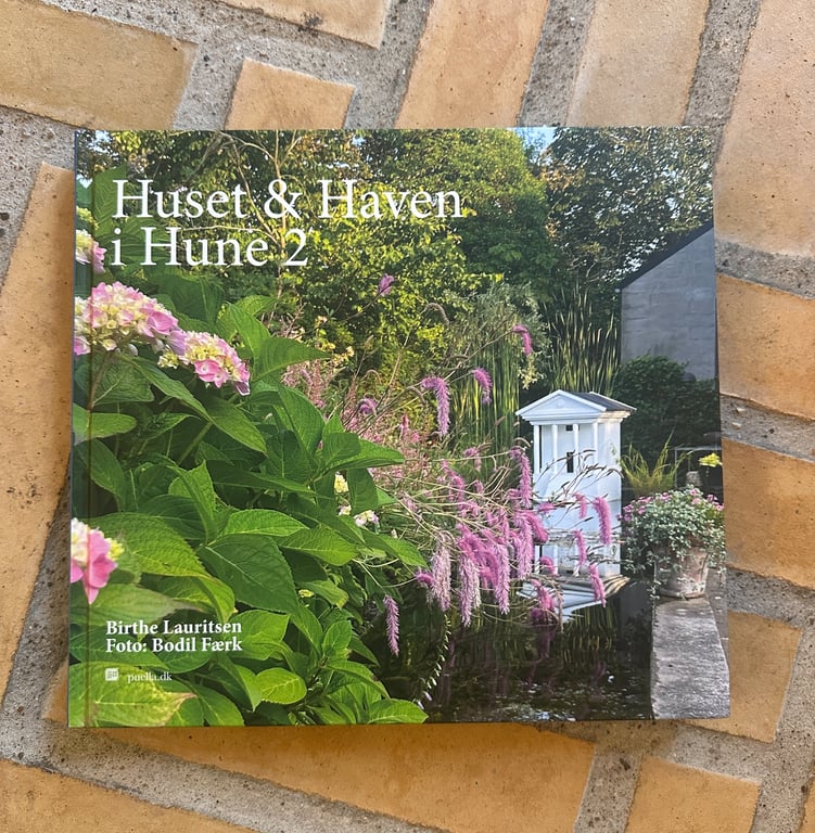 Bog med titlen "Huset & Haven i Hune 2" med et billede af en frodig have med grønne blade, pink blomster og en hvid havebænk på forsiden, lagt på flisebelagt gulv.