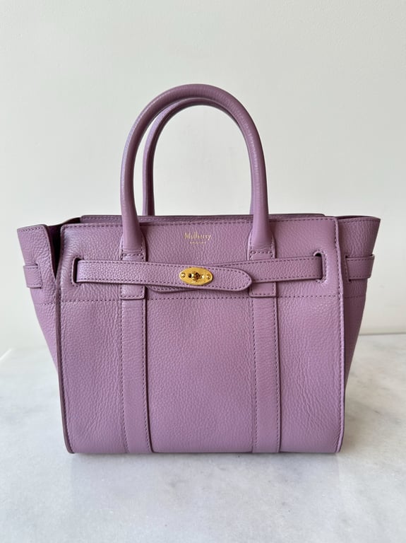 Lilla lædertaske med gyldent spænde og to håndtag, stående på hvid overflade.