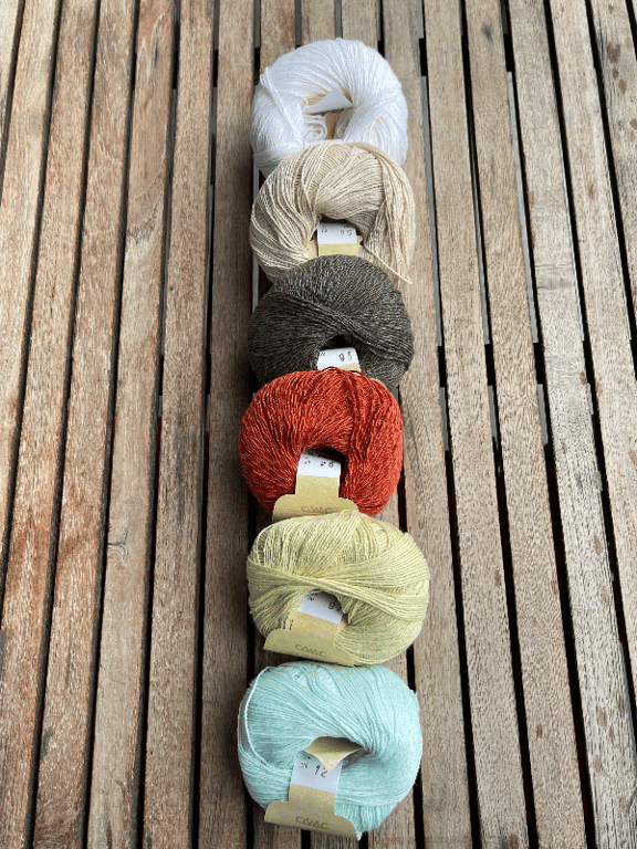 Syv farvede garnnøgler lagt i en række på et træbord, fra hvid øverst til lyseblå nederst.