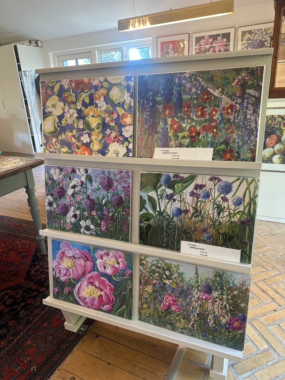 Stativ med seks farverige blomsterbilleder i et lyst rum med trægulv og tæpper.