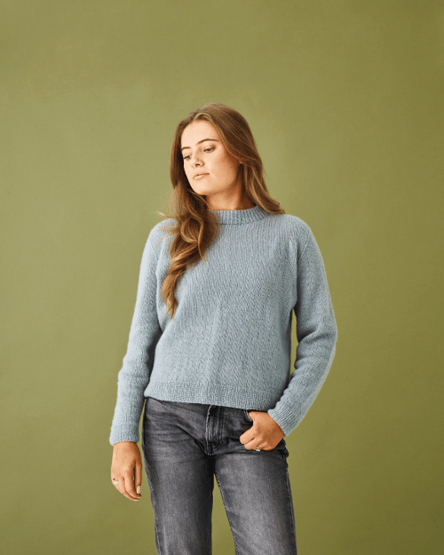 Kvinde med langt lyst hår iført lyseblå strikket sweater og mørke jeans, stående mod en grøn baggrund. (1)