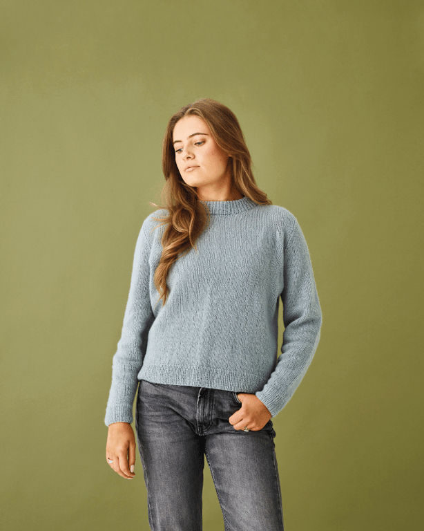 Kvinde med langt lyst hår iført lyseblå strikket sweater og mørke jeans, stående mod en grøn baggrund.