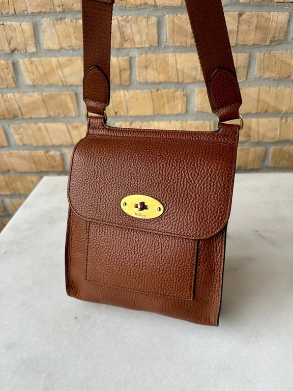 Brun læder skuldertaske med guldfarvet lukning, placeret på hvid bordplade foran en murstensvæg.