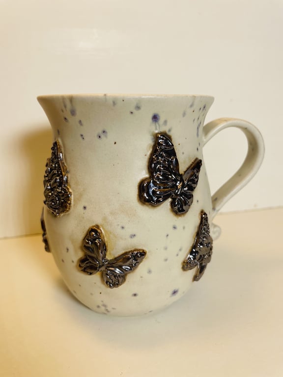 Keramikkrus med hank, dekoreret med mørke, skinnende sommerfugle på lys, priklet overflade.