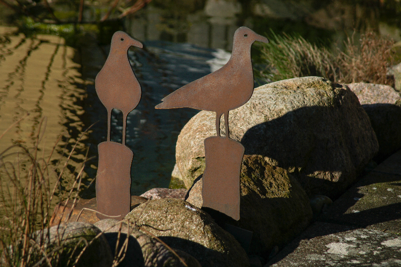 To rustne fugle skulpturer på sten ved en dam i sollys.
