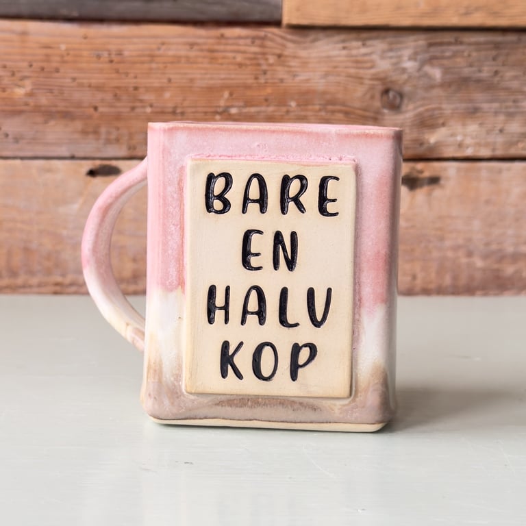 Lyserød og beige keramisk kop med teksten "Bare en halv kop" foran en træbaggrund.