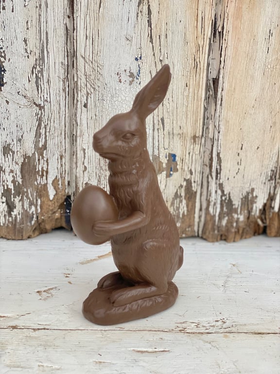 Chokoladehare, der sidder oprejst og holder et chokoladeæg, foran en rustik, afskallet træbaggrund.