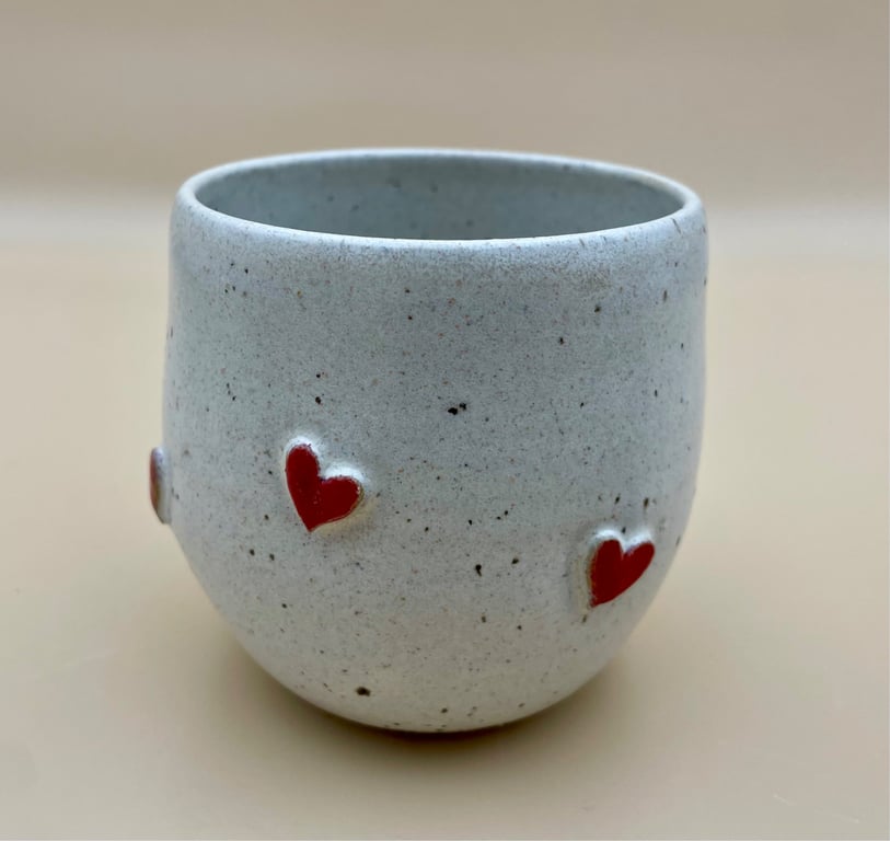 Hvid keramikkop med små røde hjerter på ydersiden, sat på en beige baggrund.