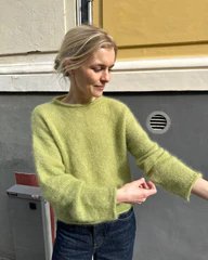 En kvinde med lyst hår er iført en grøn, strikket sweater og ruller ærmerne op, mens hun står udendørs foran en gul og grå væg. (1)