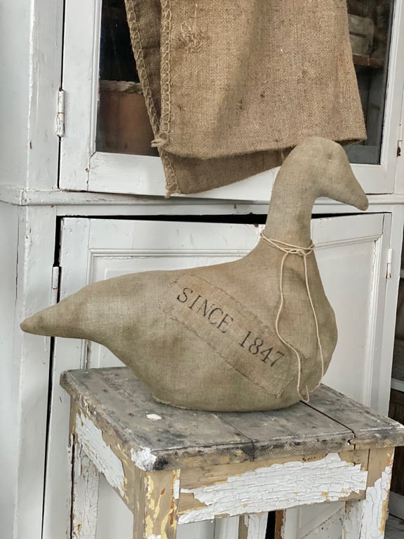 En stofdørstopper formet som en and med teksten "SINCE 1847," placeret på en slidt og afskallet træskammel foran et gammelt, hvidt træskab.