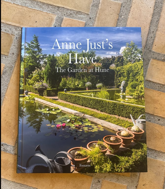 Bogomslag med titlen "Anne Just's Have - The Garden at Hune", der viser en smuk have med en dam med vandliljer, beskårne hække og potteplanter på en stenflise.