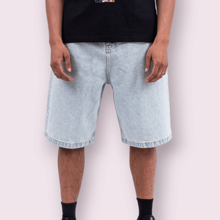 Mand iført lyseblå denimshorts, sort t-shirt og sorte sokker mod en lyserød baggrund.