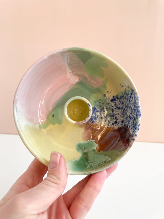 Hånd, der holder et keramisk, farverigt, rundt lille fad med en cirkulær forhøjning i midten, malet i nuancer af gul, grøn, blå, brun og lyserød.