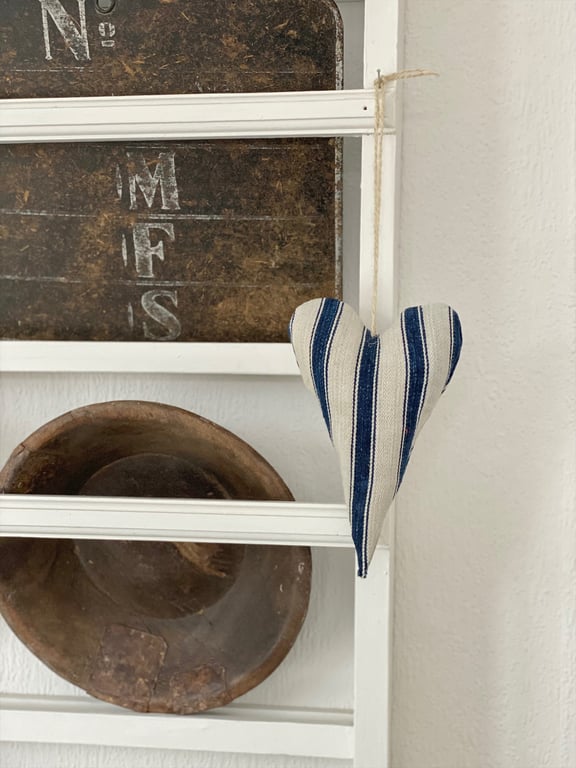 Hvid hjertedekoration med blå striber hængende på hvid ramme, baggrund med mørk rustik plade og en antik træskål.