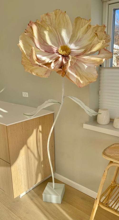 Stor, kunstig blomst med lyse blomsterblade og grønne blade, placeret på en hvid firkantet fod i et lyst rum med træmøbler og vindue. (1)