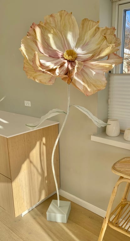 Stor, kunstig blomst med lyse blomsterblade og grønne blade, placeret på en hvid firkantet fod i et lyst rum med træmøbler og vindue.