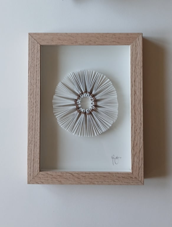 Indrammet kunstværk med et rundt, foldet papirdesign, der ligner en blomst eller sol, monteret på hvid baggrund.