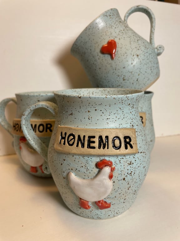 Tre lyseblå, spraglede keramikkrus med tekst "HØNEMOR" og udsmykning af en hvid høne med røde detaljer, samt et krus på skrå med et rødt hjerte.