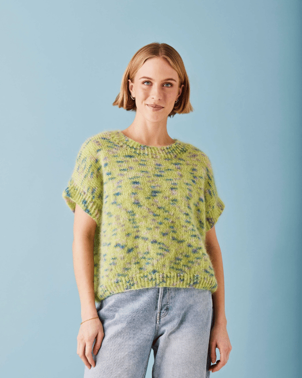 Kvinde iført kortærmet, grønmeleret striktrøje og lyseblå jeans stående foran en lyseblå baggrund.