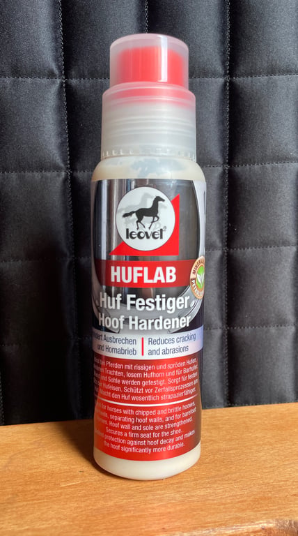 Flaske med Leovet Huflab hovforstærker til heste, placeret på en træoverflade med mørk quiltet baggrund.