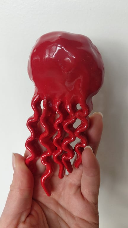 Hånd, der holder en rød, bølget blækspruttefigur i keramik mod en hvid baggrund.