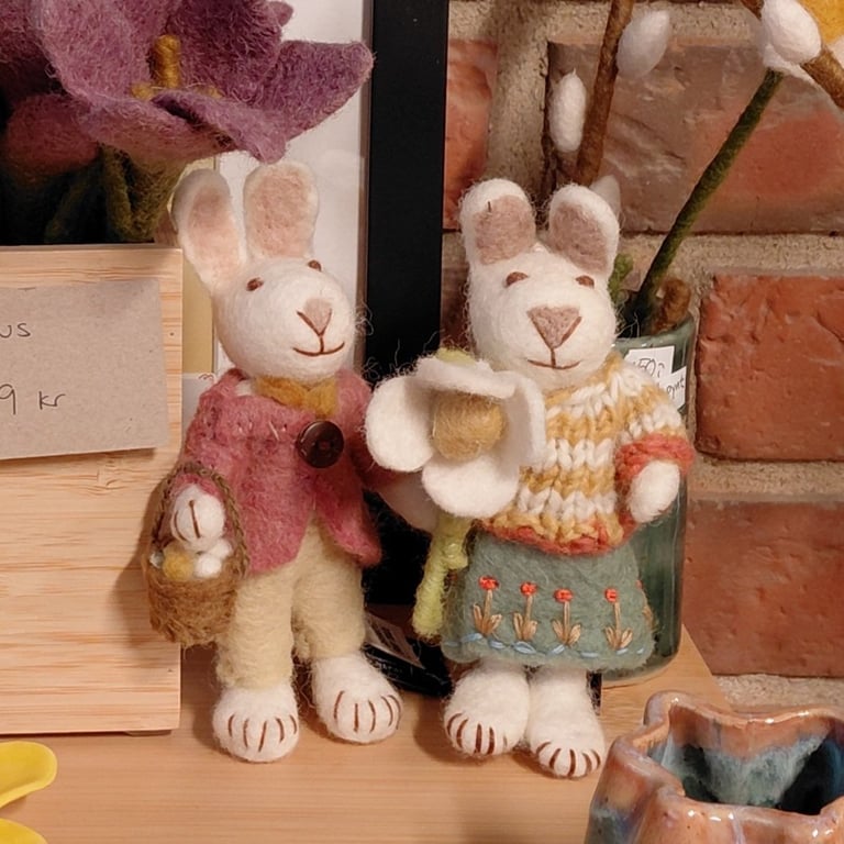 To filttede kanindukker iført farverige tøj, stående på et bord ved siden af dekorative blomster og en keramikskål.