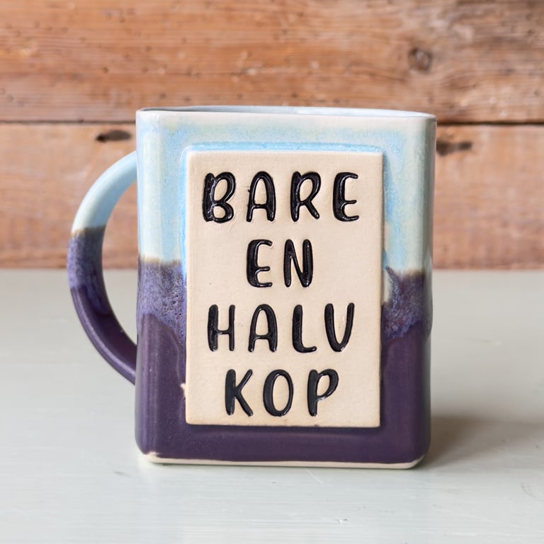 Kop i keramik med tekst "Bare en halv kop" i sort på beige firkant, farvet i blå og lilla nuancer.