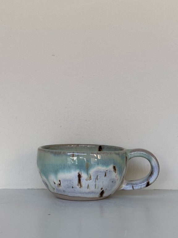 Keramikkop med blå og hvide glasurpletter og et rundt håndtag, placeret på en lys overflade mod en hvid væg.